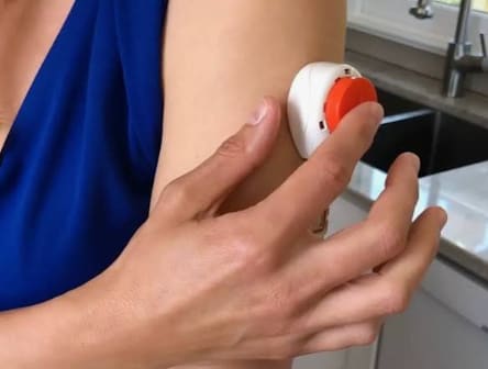 A hand pressing a small white blood collection device with an orange button against the upper arm.